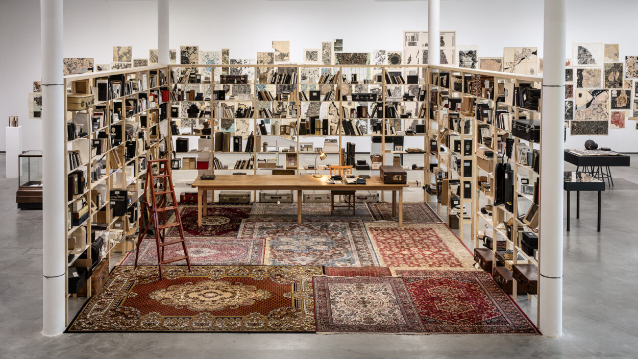 Utställningsbild med stora bokhyllor fulla av böcker och objekt. På golvet ligger orientaliska mattor. Fotografi.