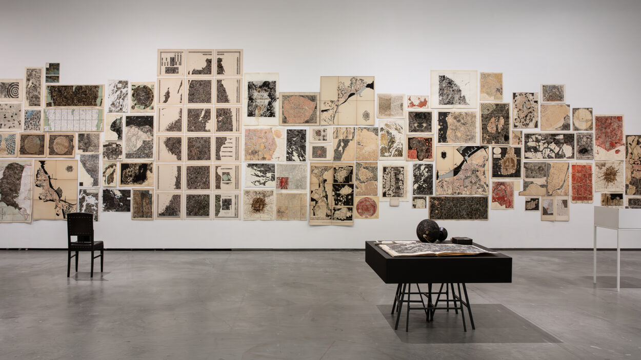 Utställningssbild med en vägg med många kartor och ett par podier på golvet framför. Fotografi