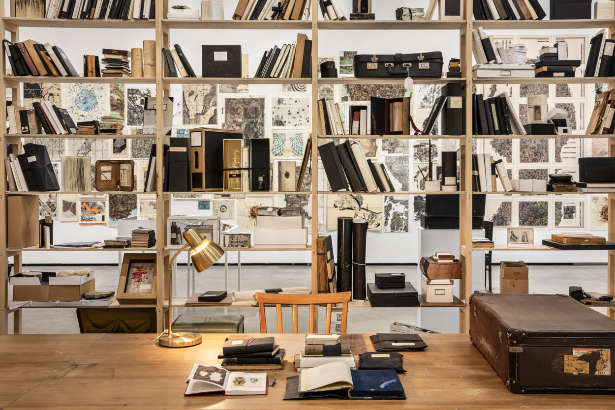 Ett skrivbord med skrivbordslampa framför en bokhylla med många olika sorters böcker. Foto.